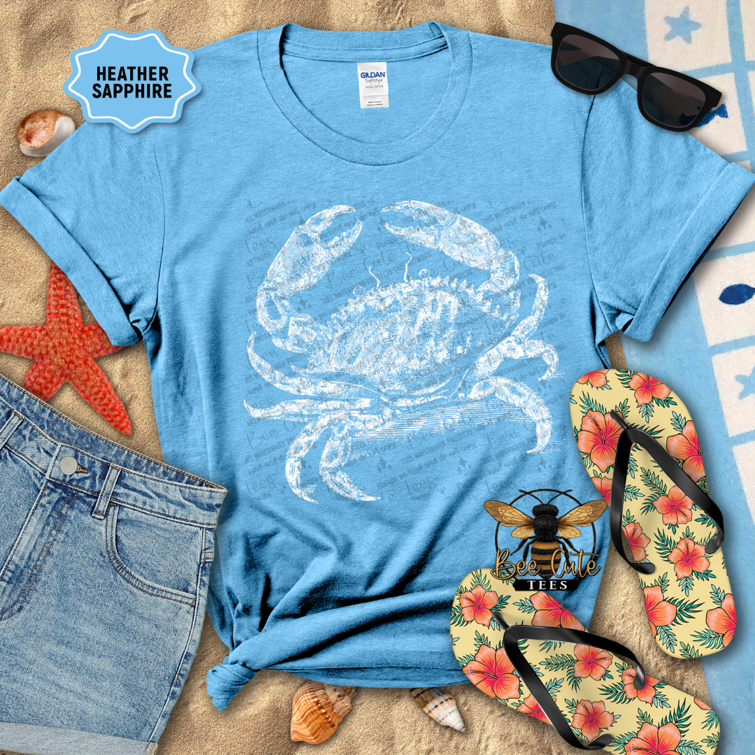 Blue t-shirt with a white crab design, sunglasses, flip-flops, and beach accessories on a sandy surface.