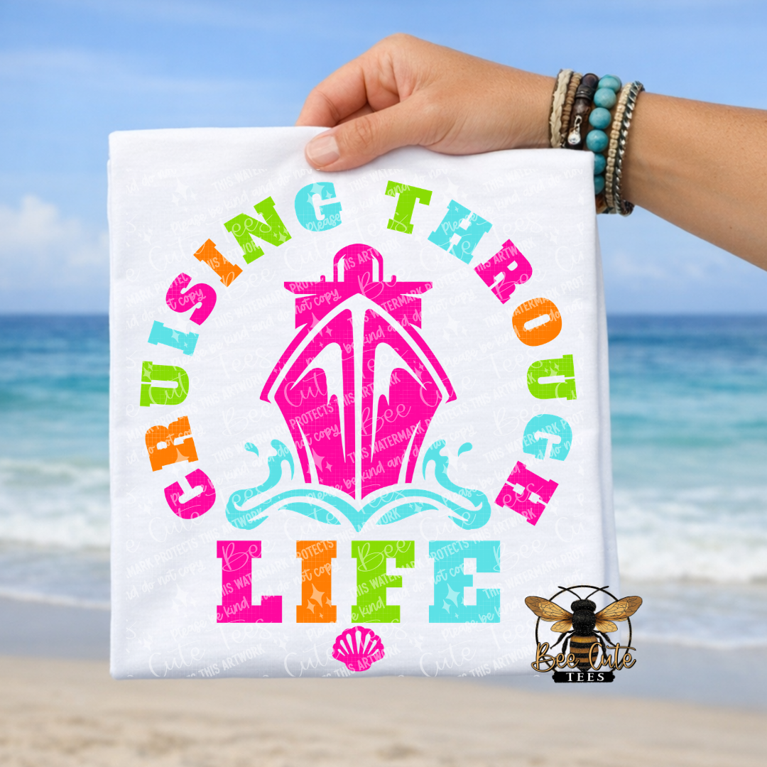 Hand holding a t-shirt with 'Cruising Through Life' text and ship graphic on a beach background