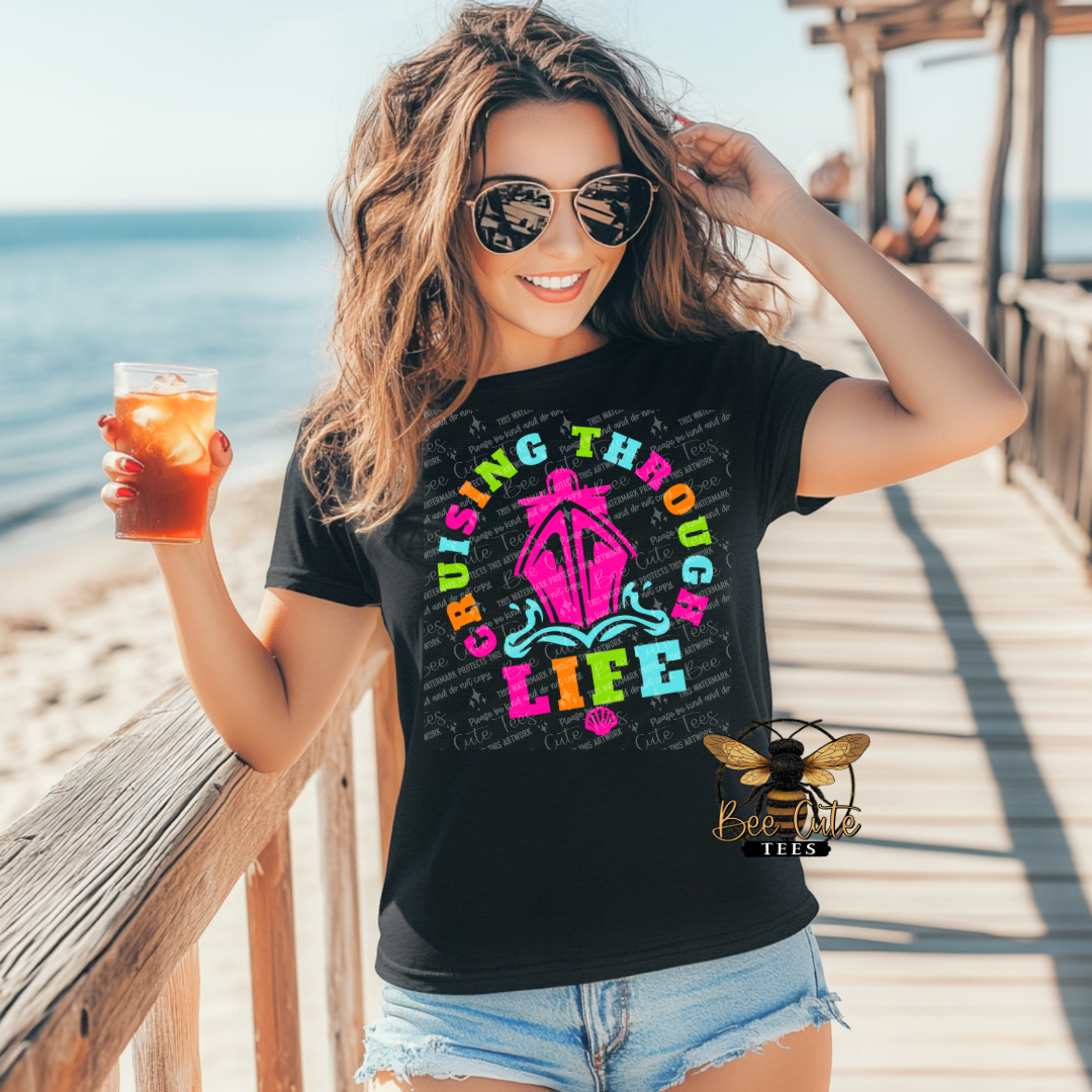 Woman on a beach holding a drink, wearing a black t-shirt with colorful text and graphics.