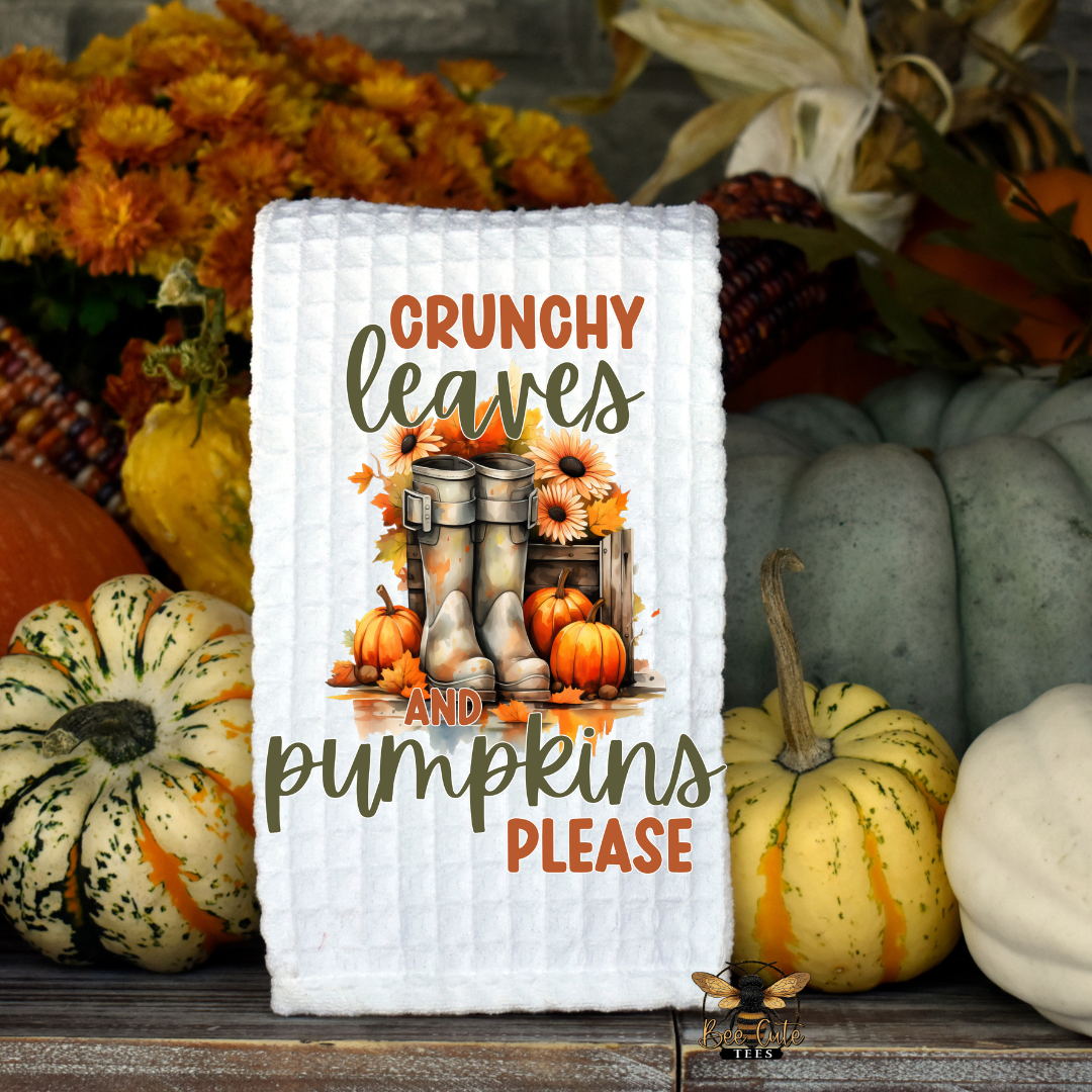 A white quilted kitchen towel with a fall-themed graphic reading 'CRUNCHY leaves AND pumpkins PLEASE' is displayed on a rustic wooden surface surrounded by pumpkins, gourds, and autumn flowers.