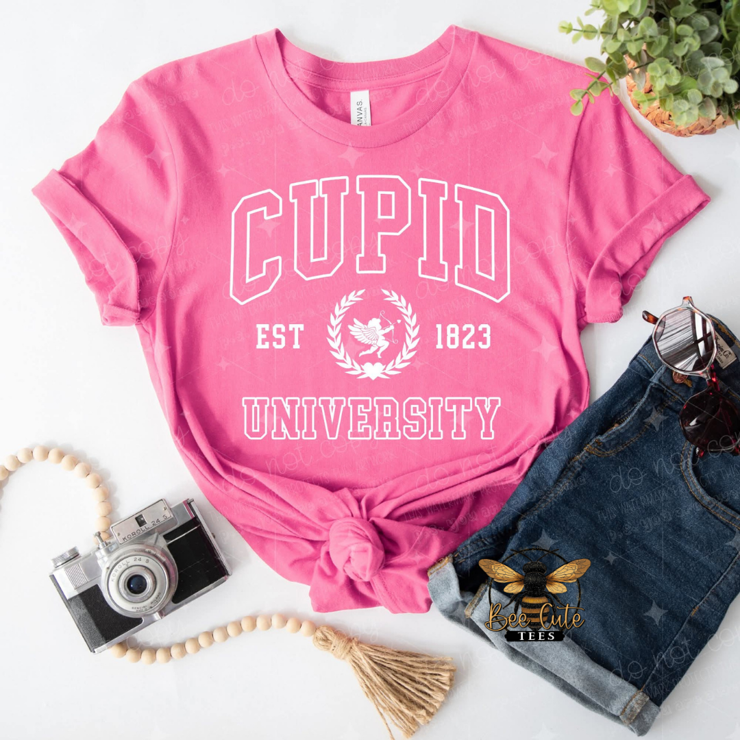 Pink t-shirt with 'Cupid University' text, camera, denim shorts, and plant on a white background