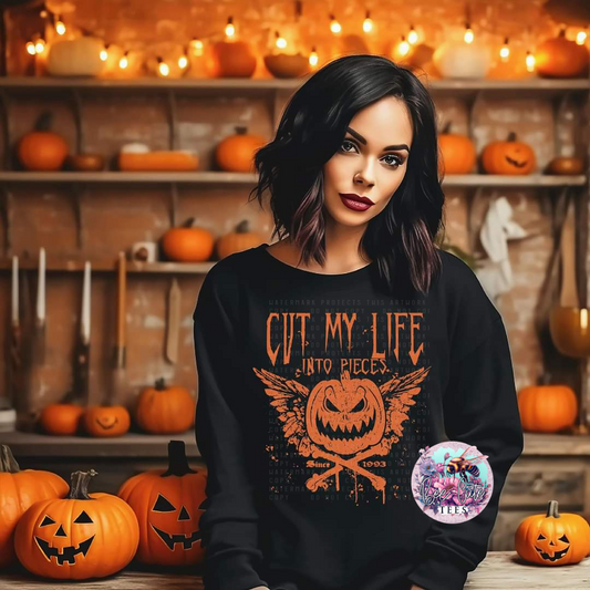 A person wearing a black sweatshirt with an orange screen print that reads 'CUT MY LIFE INTO PIECES' and features a jack o'lantern graphic, posing in a room with pumpkins and a fireplace.