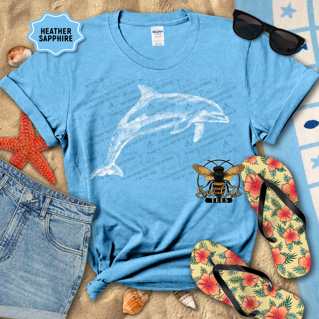Blue t-shirt with dolphin design, sunglasses, flip-flops, and denim shorts on a sandy surface.