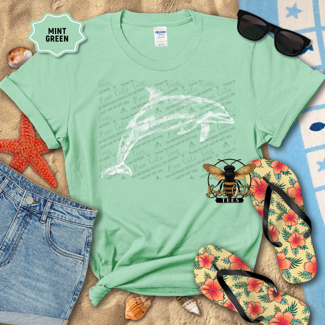 Mint green t-shirt with dolphin design, sunglasses, flip-flops, and beach accessories on sand.