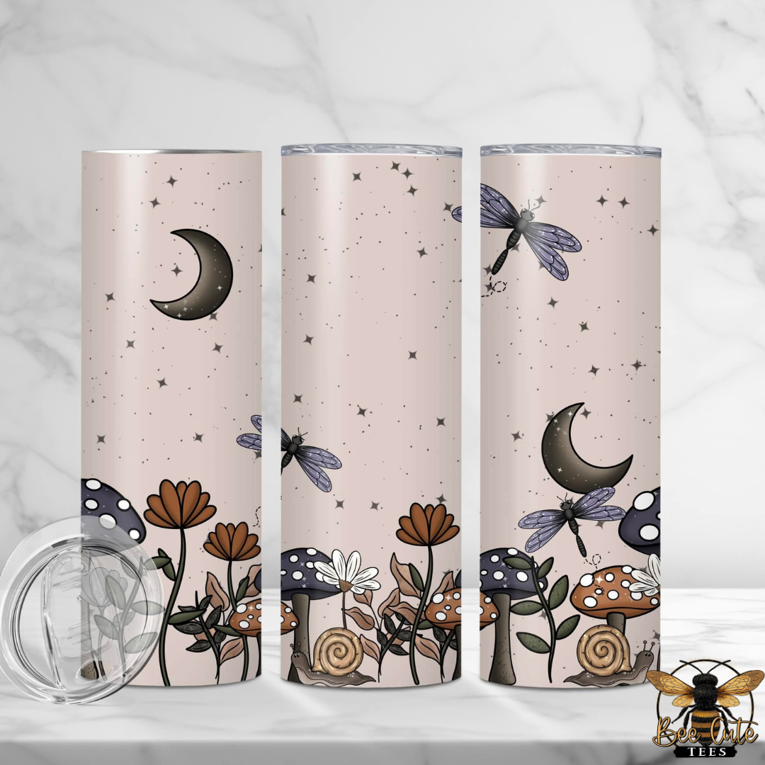 Three tall insulated tumblers with clear lids are shown on a light marble surface and background. The tumblers have a pale blush pink base with a whimsical fantasy print featuring dark gray crescent moons, blue dragonflies, brown and white mushrooms, and small stars.