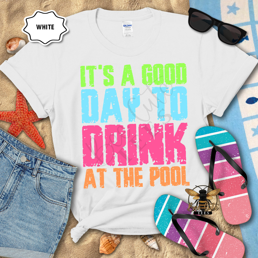 White graphic tee with colorful distressed lettering that reads “It’s a Good Day to Drink at the Pool,” styled with beach accessories including flip-flops, sunglasses, denim shorts, seashells, and a starfish.
