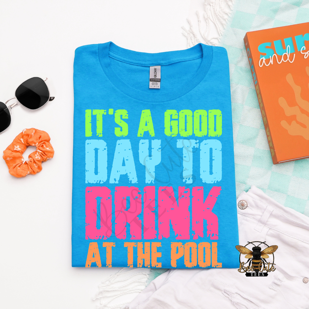 Bright blue folded graphic tee with colorful distressed lettering that reads “It’s a Good Day to Drink at the Pool,” styled with sunglasses, a scrunchie, white shorts, and summer accessories.
