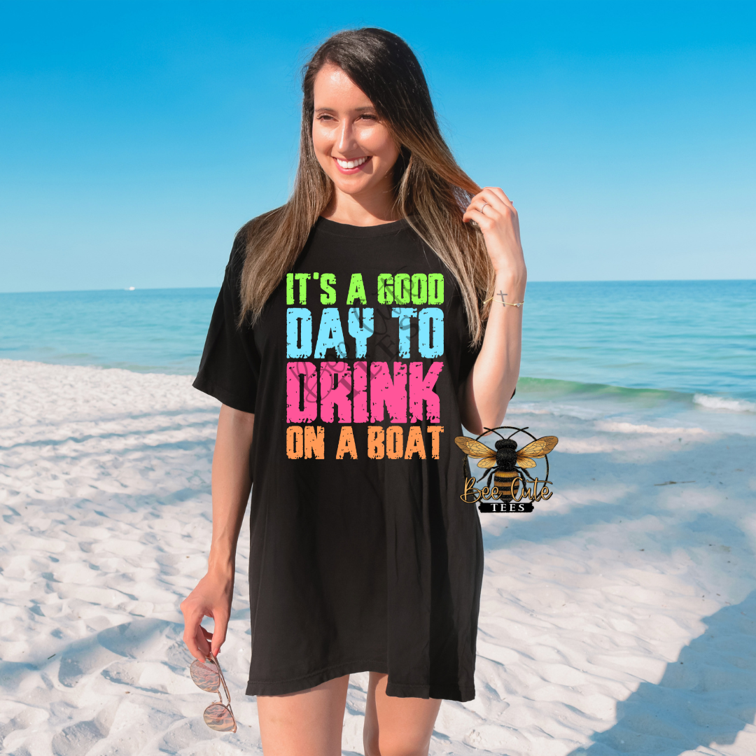 Woman standing on a sunny beach wearing an oversized black graphic tee with bright neon text that reads “It’s A Good Day To Drink On A Boat,” holding sunglasses with the ocean and white sand in the background.
