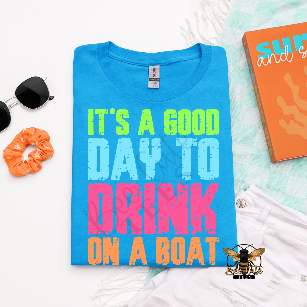 Folded bright blue graphic tee featuring colorful distressed text that reads “It’s A Good Day To Drink On A Boat,” styled with sunglasses, an orange scrunchie, white shorts, and beach accessories.
