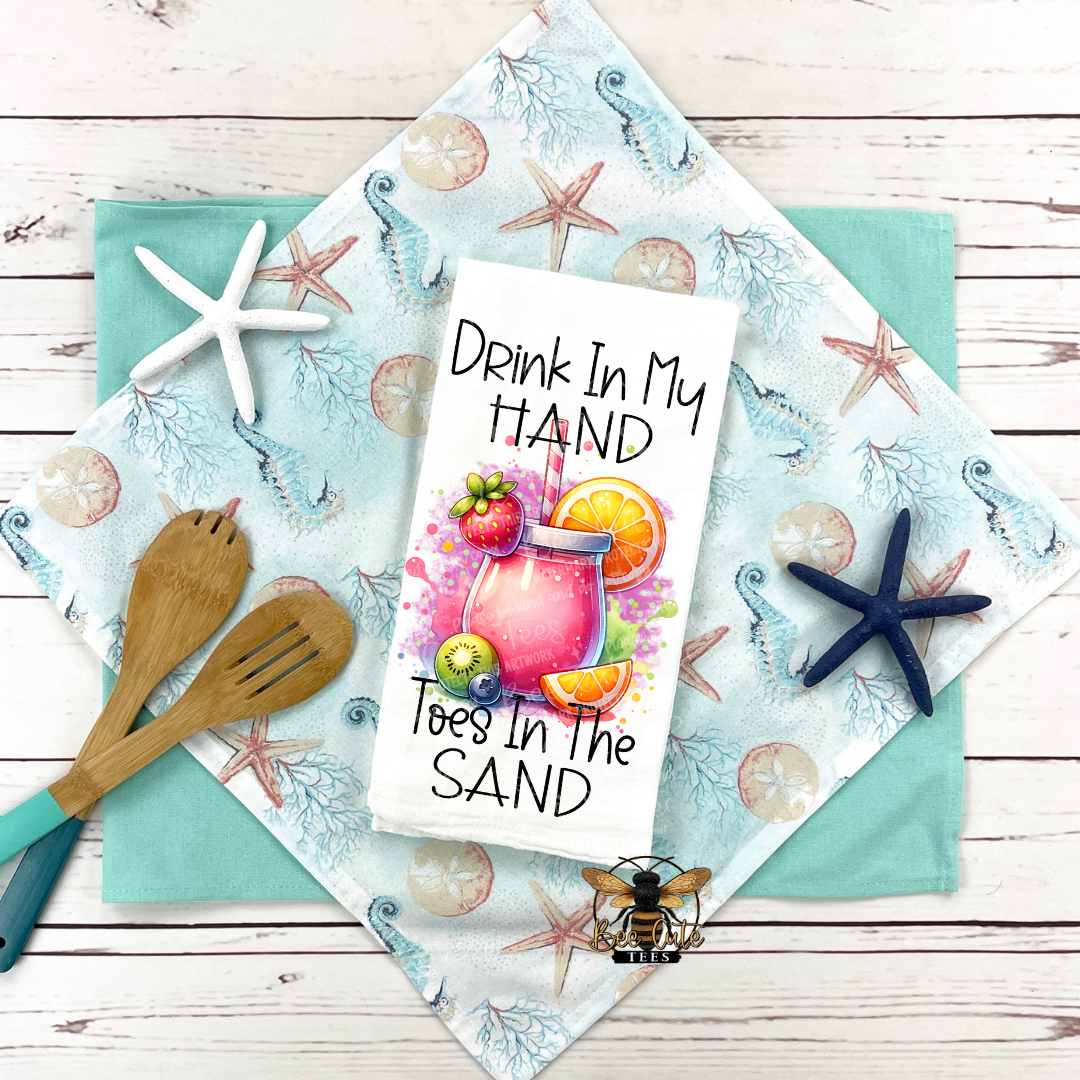 Kitchen towel with beach-themed design and text 'Drink In My Hand, Toes In The Sand' on a wooden surface.
