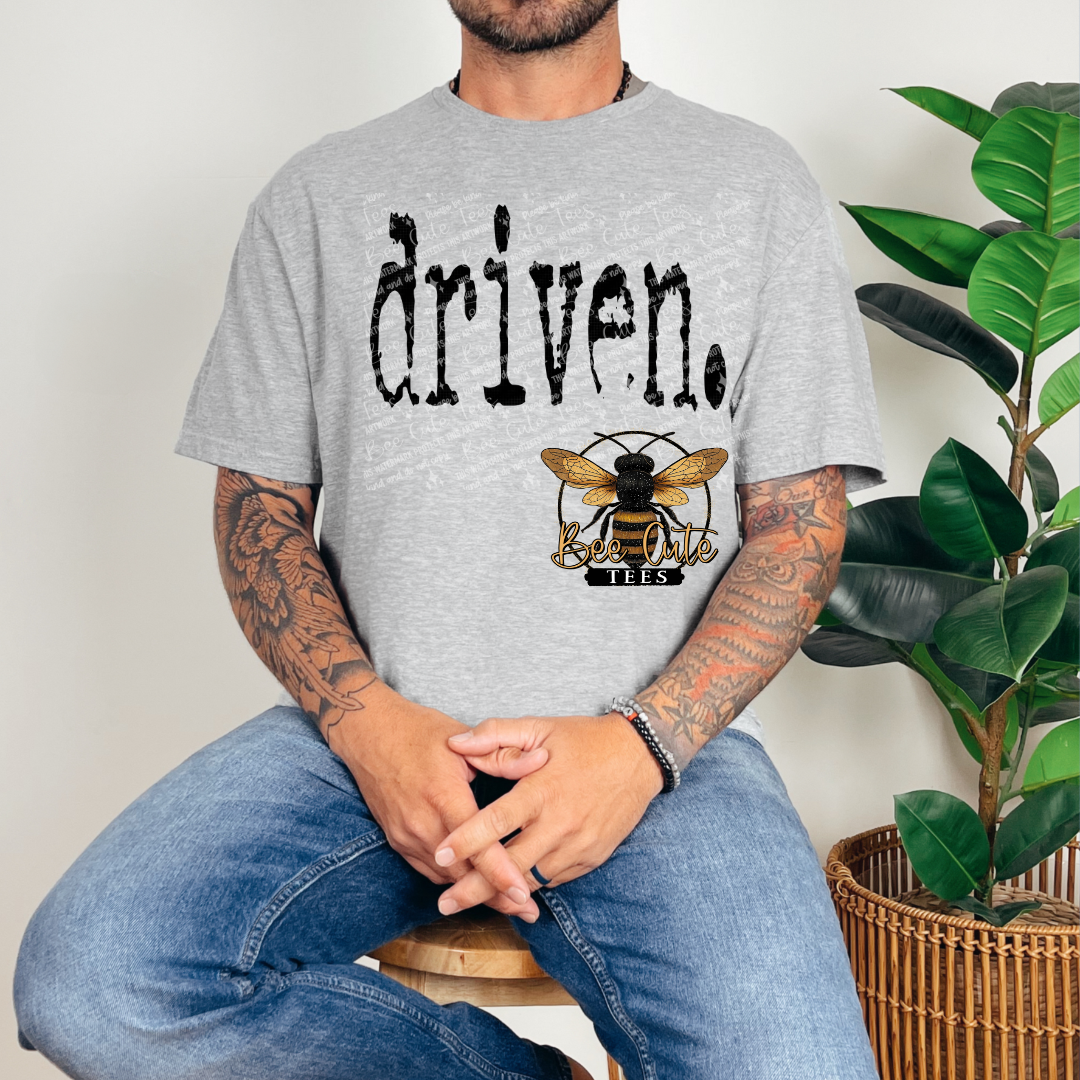 Person wearing a gray t-shirt with 'driven' and a bee logo, sitting next to a plant.