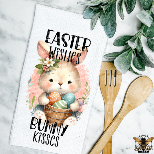 Decorative towel with Easter design featuring a bunny and text, placed on a marble surface with wooden utensils and greenery.