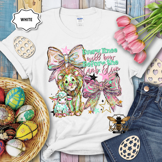 White t-shirt with colorful design featuring a lion, sheep, and decorative elements on a wooden surface with Easter decorations.
