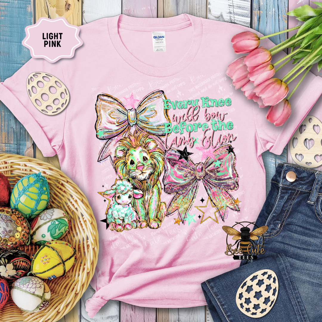 Pink t-shirt with a colorful design featuring a lion, sheep, and decorative elements on a wooden surface with Easter decorations.