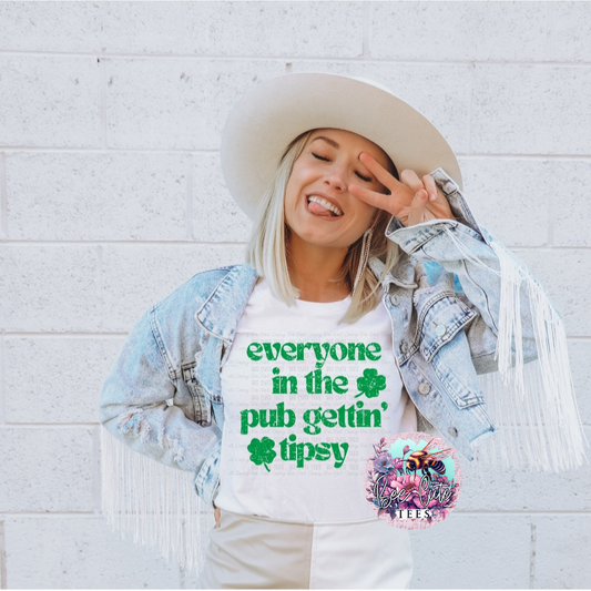 A person wearing a white hat and a short sleeve green t-shirt with the text 'EVERYONE IN THE PUB GETTING TIPSY' and a shamrock symbol on it, standing against a white wall with fringes in the background.