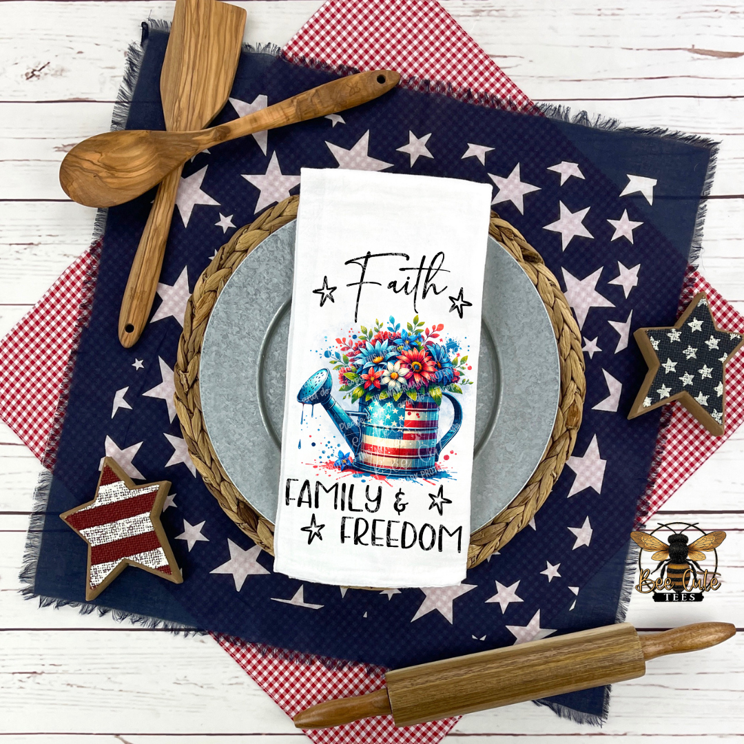 White kitchen towel displayed on a patriotic table setting, featuring a red, white, and blue watering can filled with flowers and the phrase “Faith Family & Freedom,” surrounded by star-patterned linens, wooden utensils, and rustic decor.
