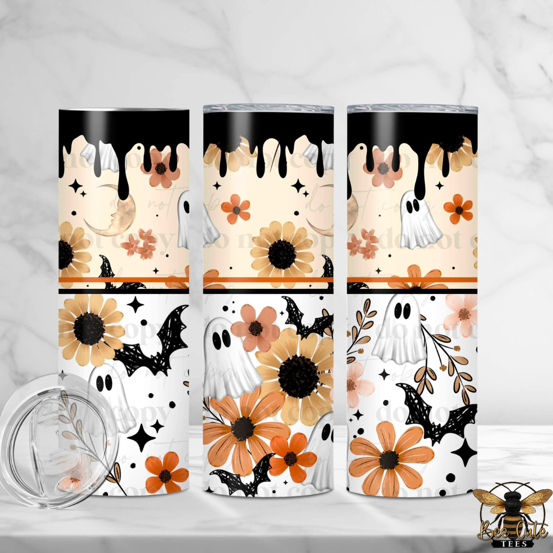Three slim stainless steel tumblers with a Halloween-themed print are displayed on a white marble surface with a white marble background. The print features a cream base with black dripping paint at the top, orange and beige sunflowers, white ghosts, black bats, and small orange flowers.