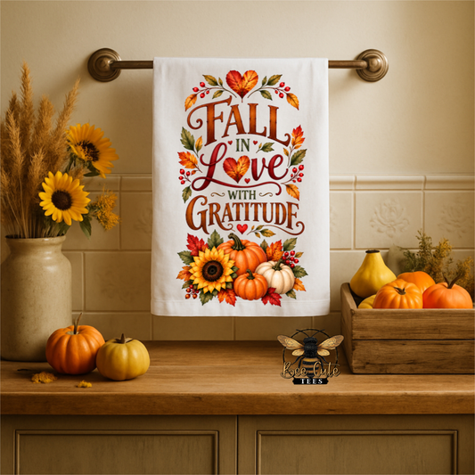 A white kitchen towel with a fall-themed graphic reading 'FALL IN Love WITH GRATITUDE' hangs on a brass towel bar. The towel features orange and brown lettering, autumn leaves, sunflowers, and pumpkins.