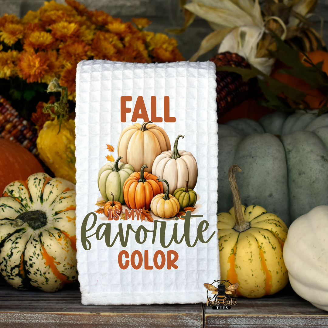 A white waffle-knit kitchen towel with a fall-themed graphic reading 'FALL favorite COLOR' and showing illustrated pumpkins, styled on a wooden surface with real pumpkins and autumn flowers in the background.