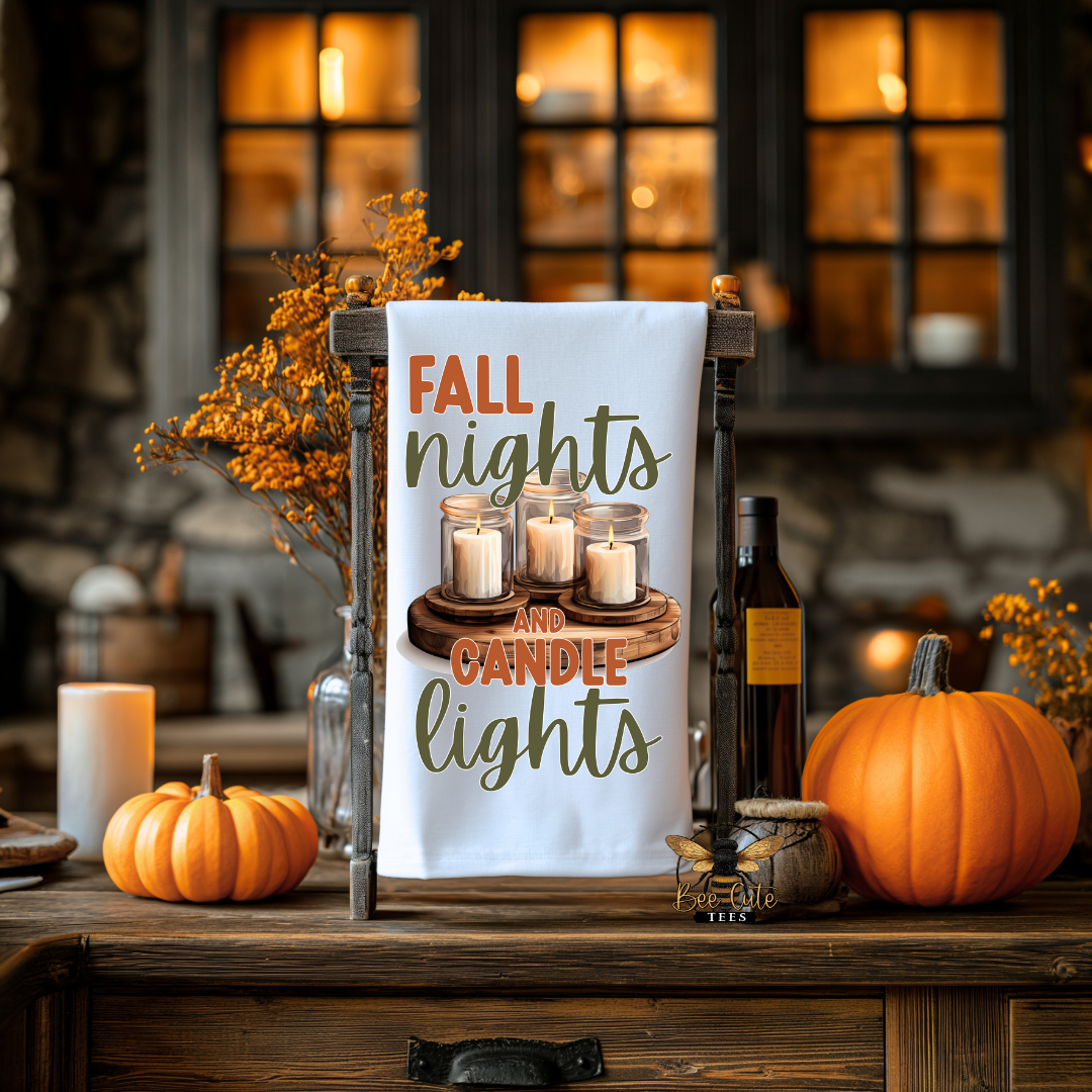 A white decorative tea towel hanging in a wooden stand on a rustic wooden table, printed with the phrase “FALL nights and CANDLE lights” in orange and green lettering above an illustration of three lit candles on a wooden tray. The scene is styled for fall with pumpkins, a candle, a bottle, and dried orange foliage in a warmly lit indoor setting.