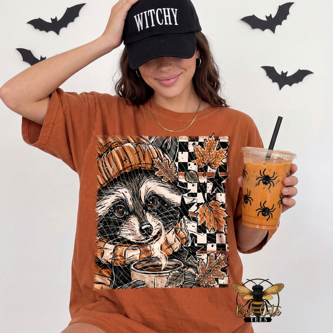 Woman wearing a rust-orange graphic tee featuring a raccoon with pumpkins, fall leaves, stars, and a checkerboard background, paired with a black “Witchy” hat and holding an iced drink decorated with spiders.