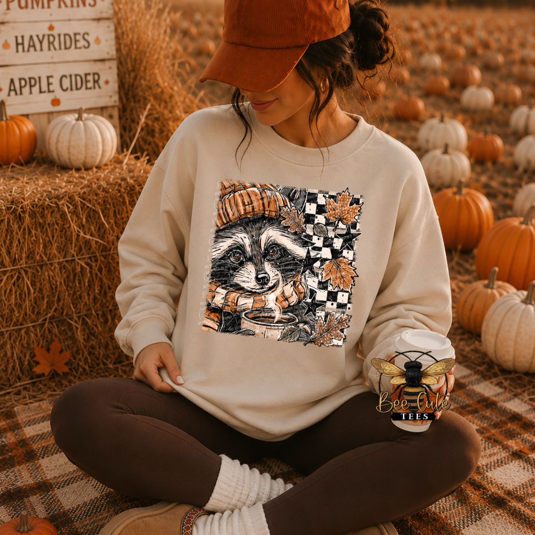 A woman sits cross-legged on a plaid blanket in a fall-themed pumpkin yard, wearing a cream sweatshirt with a large raccoon graphic and an orange cap. The sweatshirt features a detailed illustration of a raccoon wearing an orange beanie and scarf, set against a black-and-white checkerboard pattern with orange leaves.