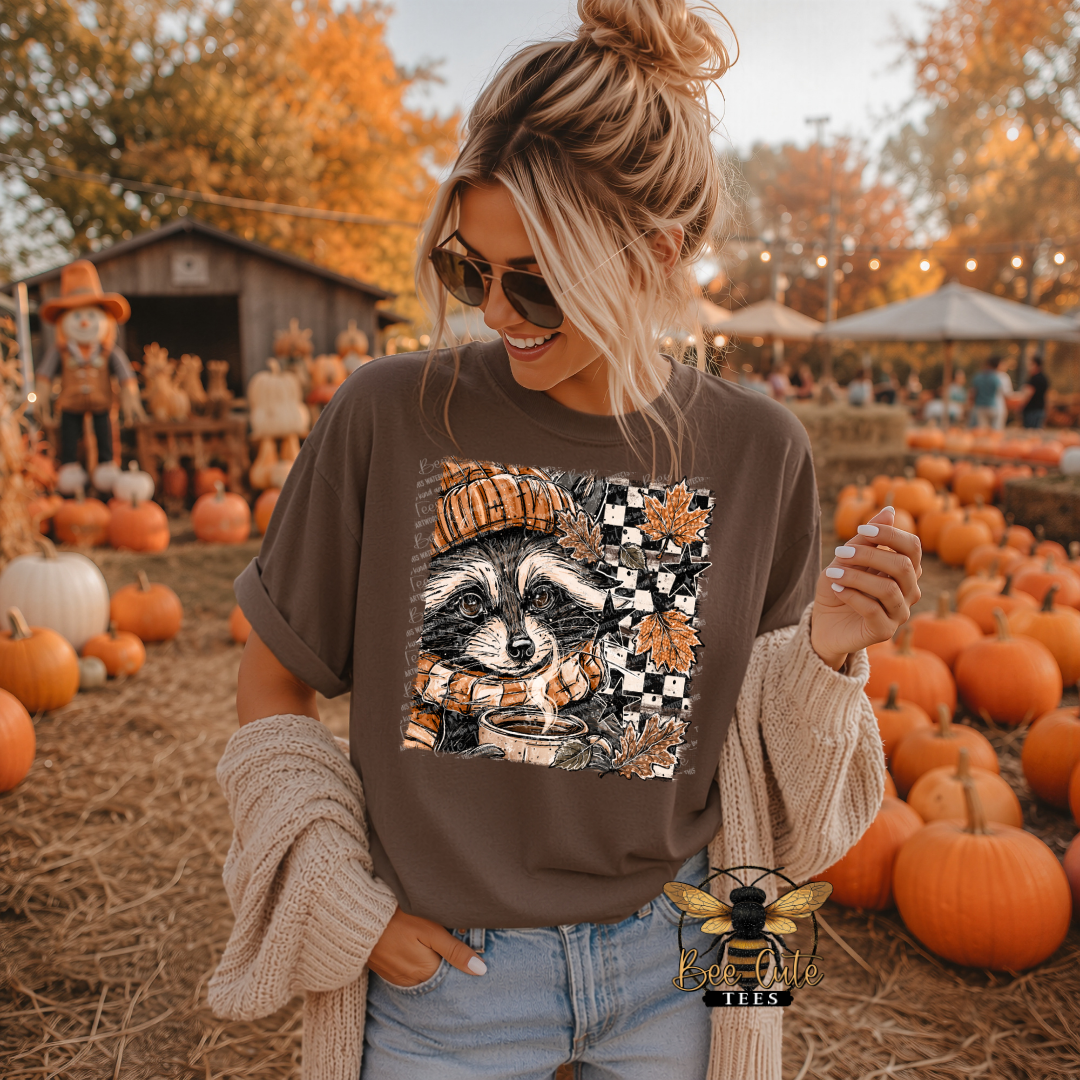 A woman wearing a brown graphic t-shirt with a raccoon design, standing in a pumpkin patch. The t-shirt features a square graphic of a raccoon wearing a striped beanie and a scarf, surrounded by autumn leaves and a checkered pattern.