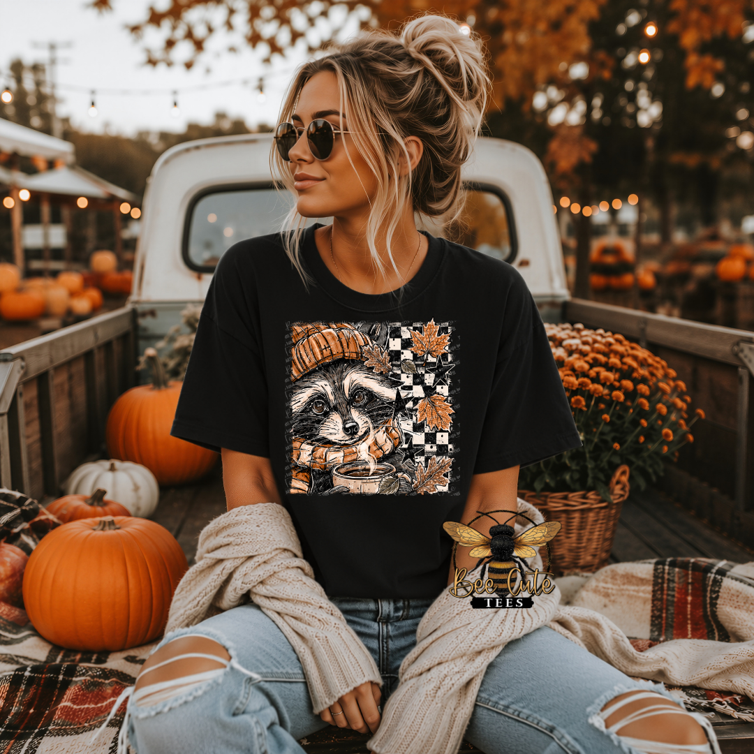 A woman wearing a black graphic t-shirt with a raccoon and autumn design, sitting in the bed of a pickup truck surrounded by pumpkins, fall foliage, and blankets.