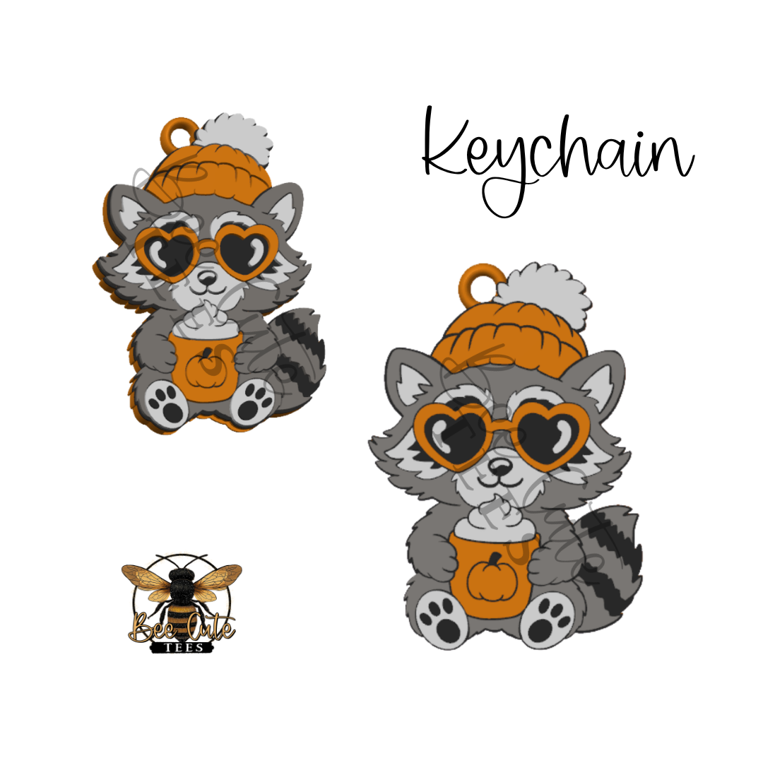 A keychain charm featuring a cartoon raccoon wearing an orange knit beanie with a white pom-pom, heart-shaped sunglasses, and holding a pumpkin cup with whipped cream. The image shows two views of the keychain, a small bee logo  at the bottom left, and the word 'Keychain' at the top right, all on a plain white background.