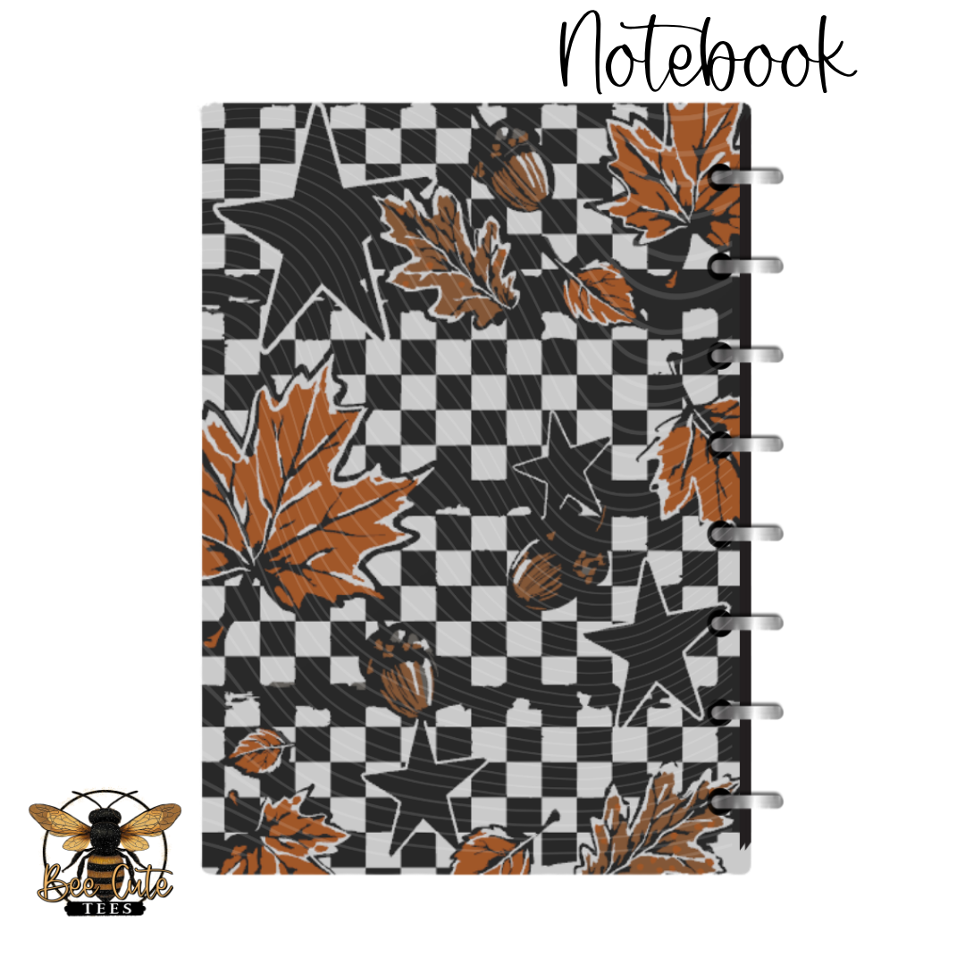 A spiral-bound notebook with a black and white checkerboard pattern, decorated with orange autumn leaves, black stars, and acorns. The word 'Notebook' is written in black script at the top right, and a bee logo is in the bottom left corner, all on a plain white background.