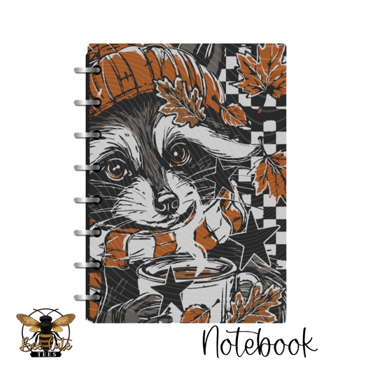 A spiral-bound notebook featuring a stylized raccoon illustration with autumn leaves, stars, and a checkered background. The word 'Notebook' appears below the product, and a bee logo is shown in the lower-left corner on a white background.