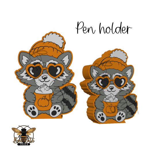 Two cute raccoon pen holders shown on a white background, each wearing an orange knit hat with a white pom-pom and heart-shaped sunglasses while holding an orange pumpkin cup. The text 'Pen holder' appears above, and a bee logo is in the lower left corner.