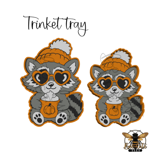 Two cartoon raccoon characters wearing orange knit beanies and heart-shaped sunglasses, each holding a small pumpkin cup, shown on a white background with the text 'Trinket tray' and a bee logo at the bottom right.