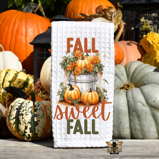 A white waffle-weave kitchen towel with a fall harvest illustration stands on a wooden surface surrounded by real pumpkins and gourds outdoors. The towel reads “FALL sweet FALL” with pumpkins, gourds, and orange flowers in a bucket.