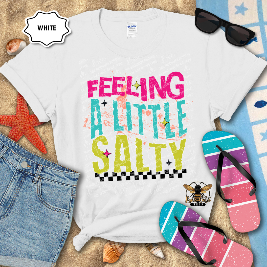 White t-shirt with 'Feeling a Little Salty' text, sunglasses, flip-flops, and denim shorts on a sandy background.