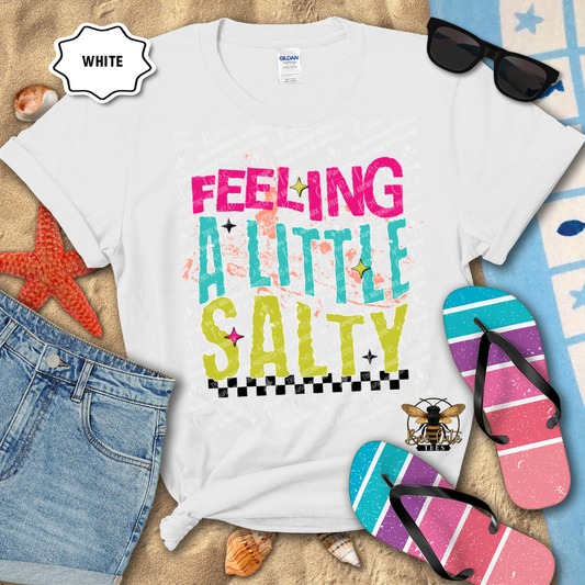 White t-shirt with 'Feeling a Little Salty' text, sunglasses, flip-flops, and denim shorts on a sandy background.