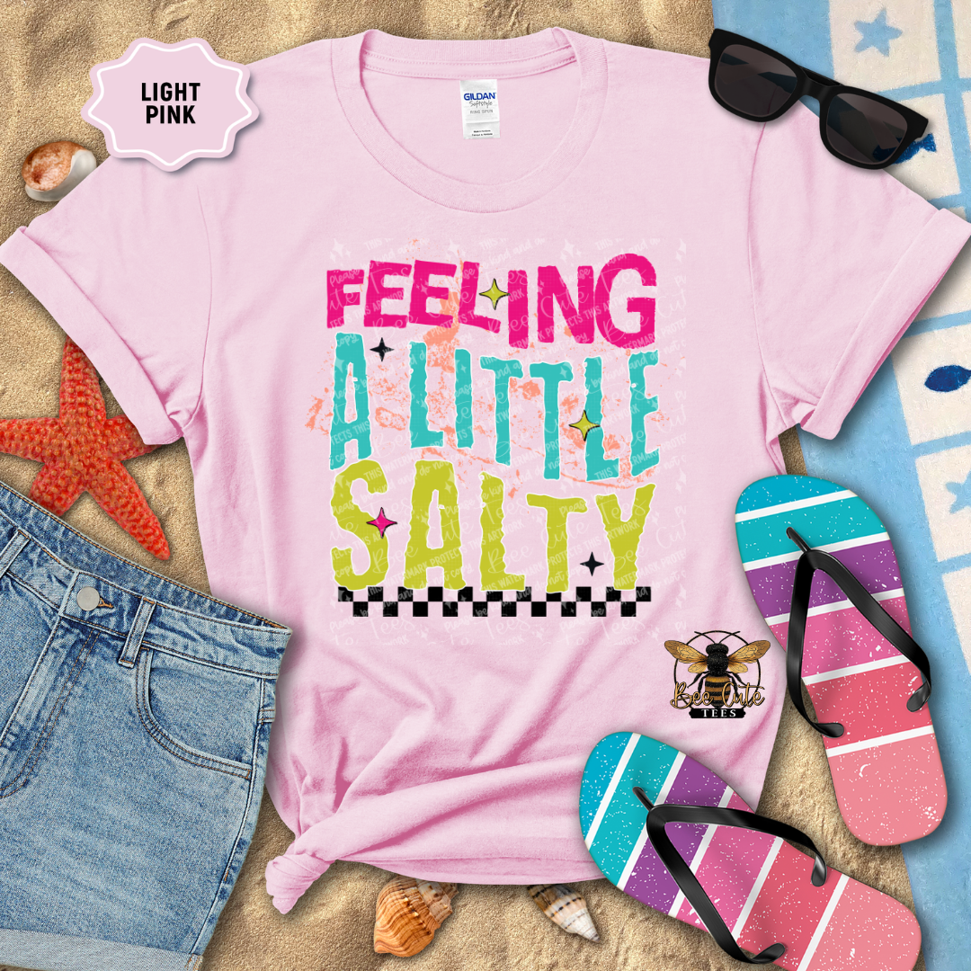 Light pink t-shirt with 'Feeling a Little Salty' text, sunglasses, denim shorts, and beach accessories on sand.