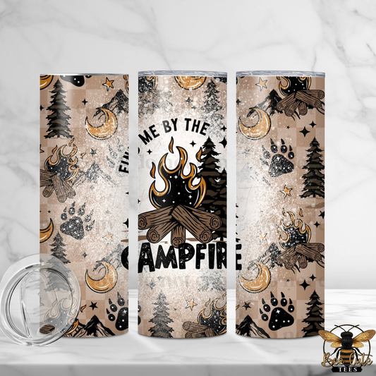Three insulated tumblers with a rustic campfire-themed wrap design, shown on a white marble surface with a clear lid in the foreground. The design features a beige distressed background with black and brown campfire, trees, moon, and paw prints, with the text 'WELCOME BY THE CAMPFIRE' on the center tumbler.