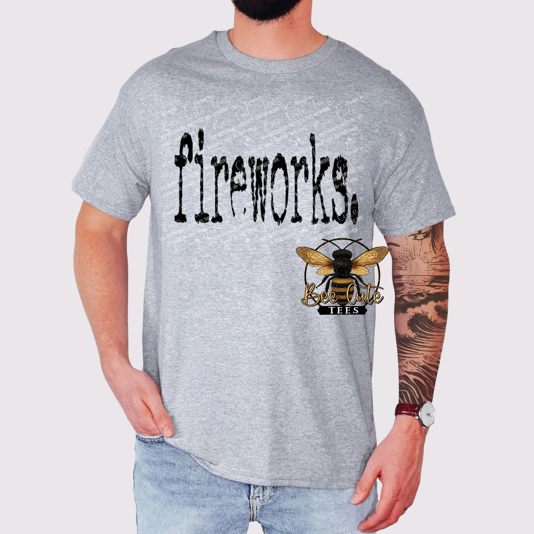 Gray graphic t-shirt with distressed black text reading “fireworks,” worn by a man with a tattooed arm, styled with jeans and a watch, featuring a Bee Cute Tees logo.
