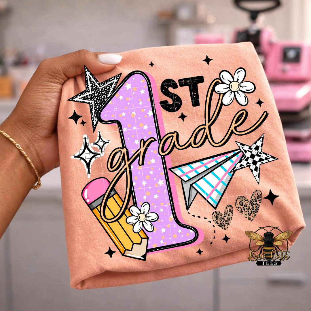 Hand holding an orange t-shirt with '1st Grade' design featuring stars, flowers, and a pencil.