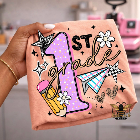 Hand holding an orange t-shirt with '1st Grade' design featuring stars, flowers, and a pencil.