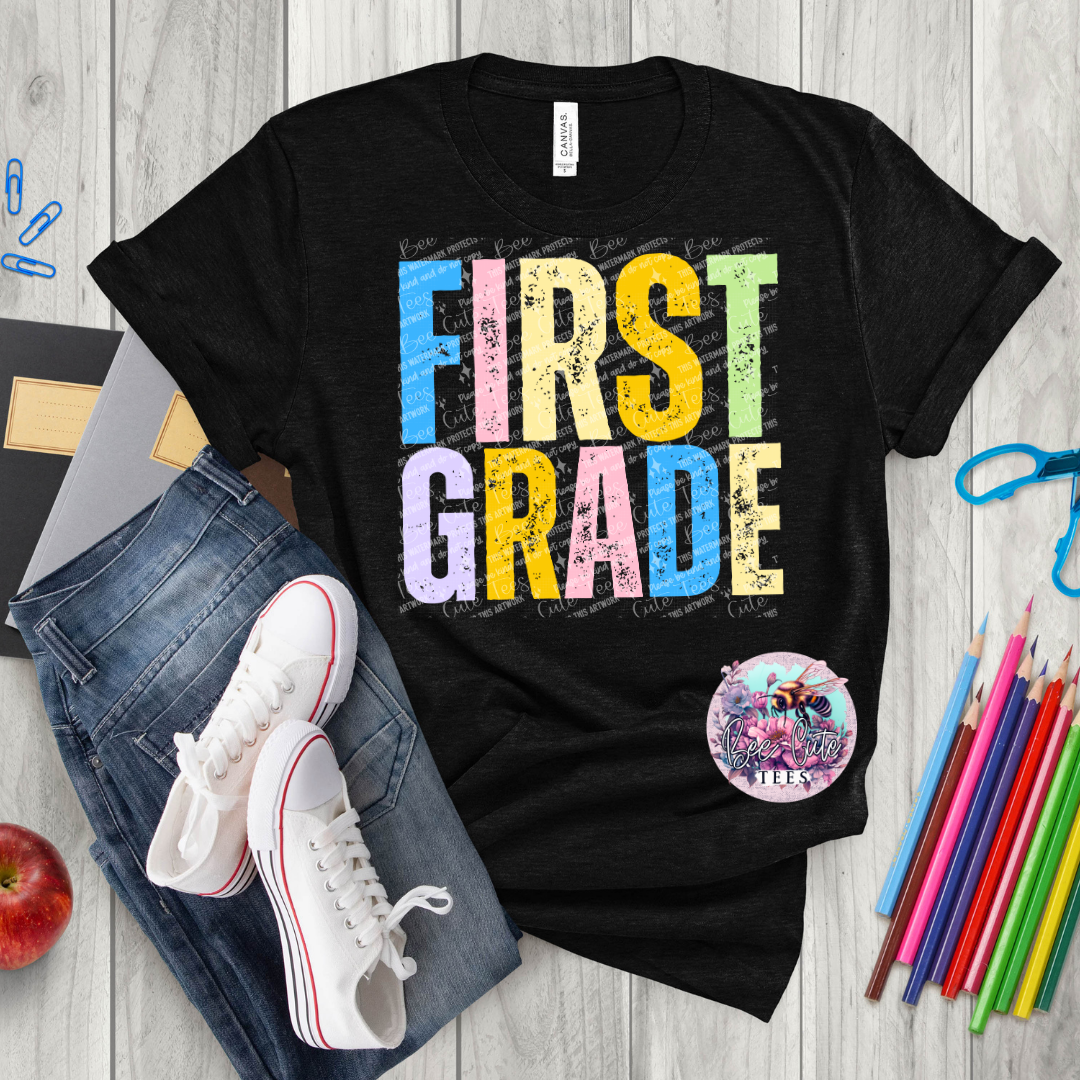Black t-shirt with 'FIRST GRADE' in colorful letters on a wooden surface with jeans, sneakers, and school supplies.