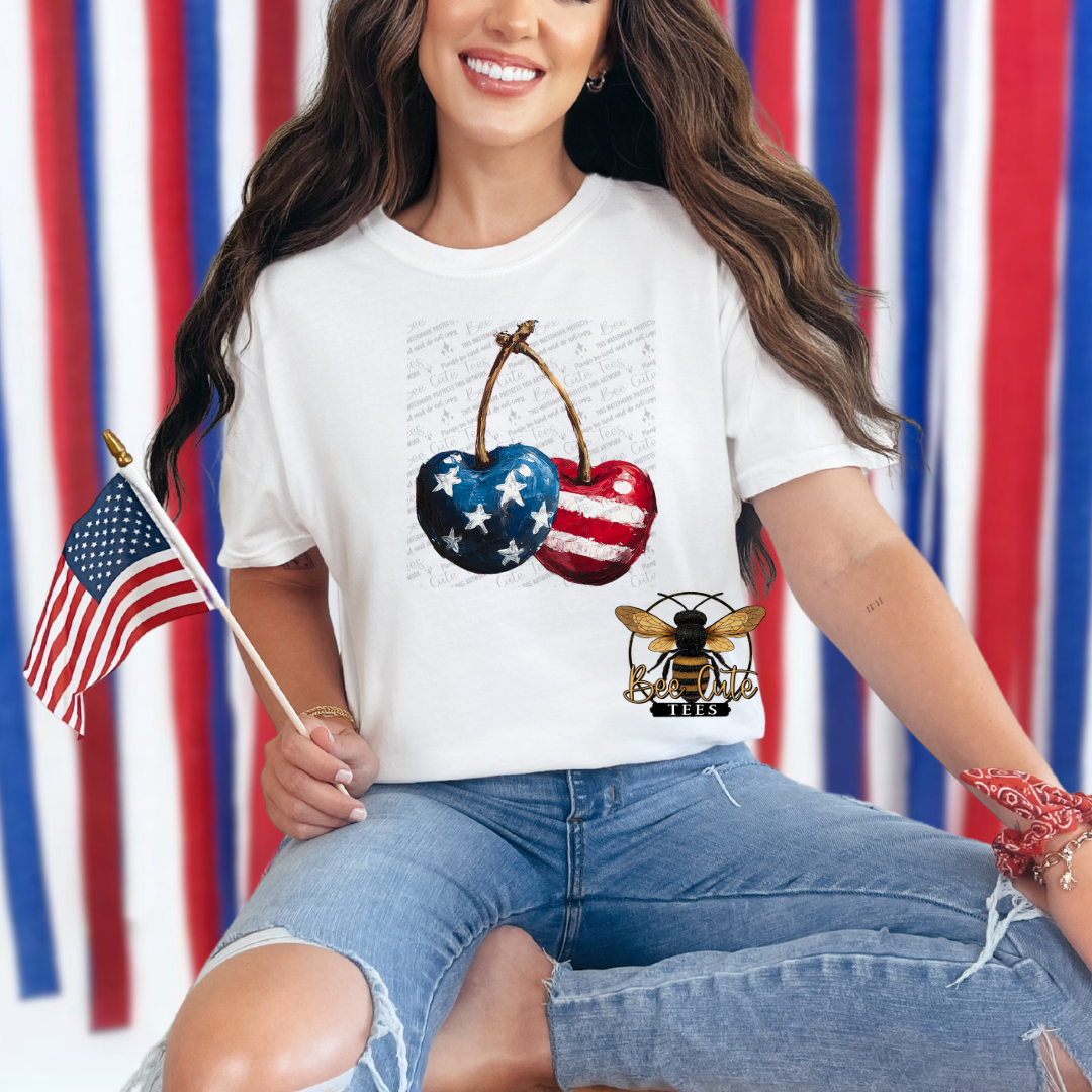 A person holding a small American flag wearing a white short-sleeve t-shirt with a graphic of two cherries, one red and one white striped, against a striped red and blue background.
