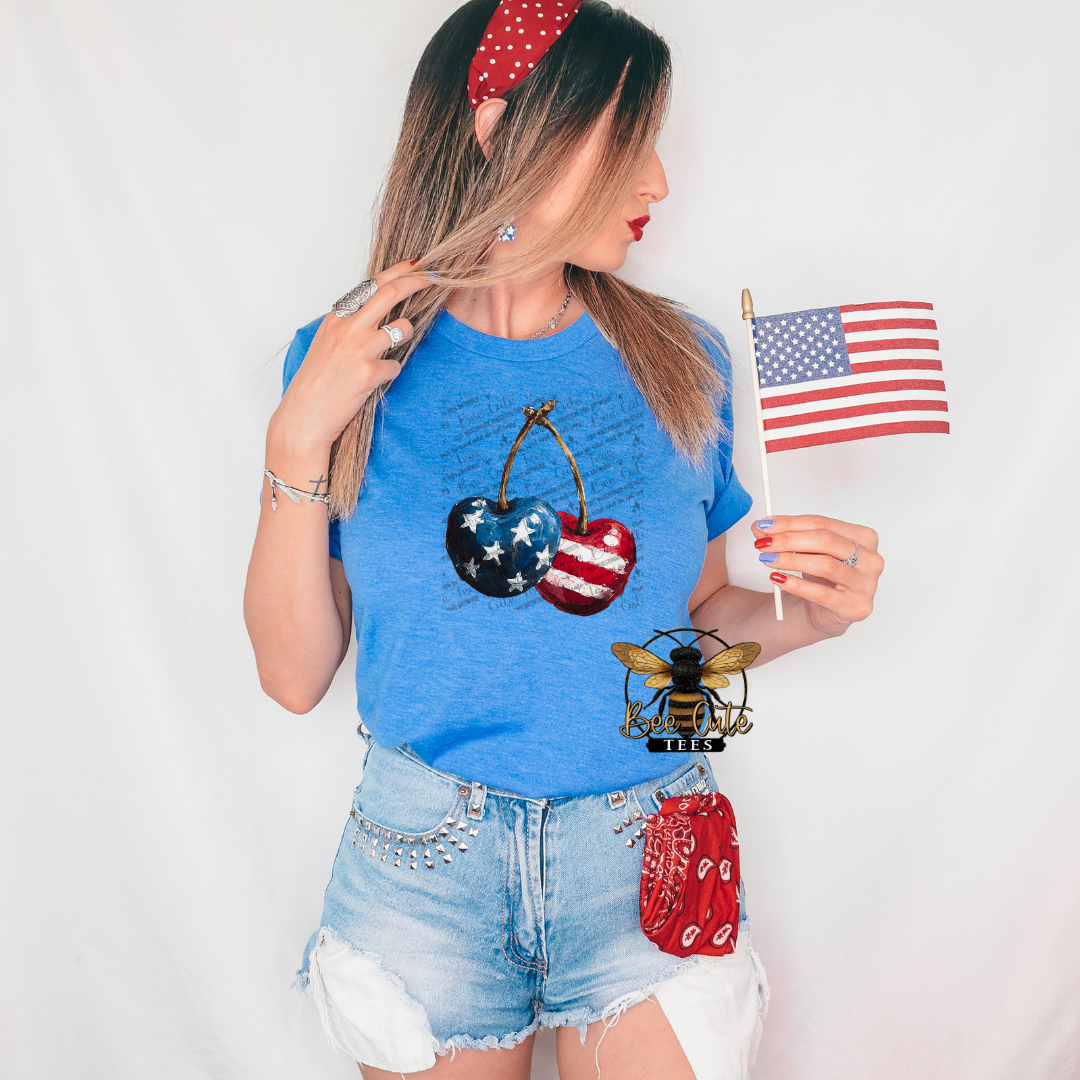 Woman wearing a heather royal blue patriotic graphic tee featuring hand-painted style cherries decorated with American flag stars and stripes, paired with distressed denim shorts and red, white, and blue accessories while holding a small American flag.
