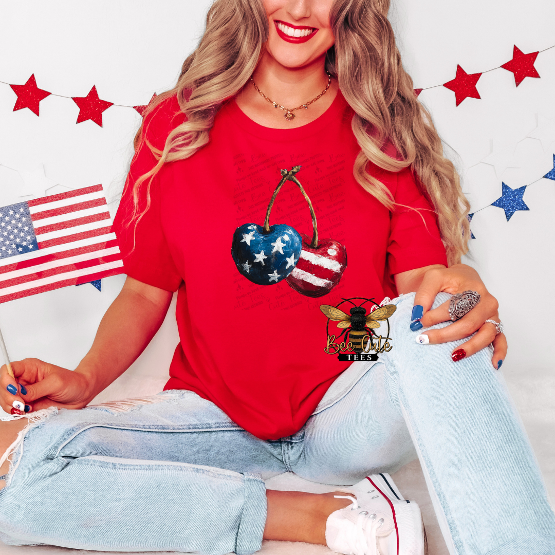 Woman wearing a bright red patriotic graphic tee featuring hand-painted style cherries decorated with American flag stars and stripes, paired with light-wash jeans and patriotic accessories against a festive red, white, and blue backdrop.

