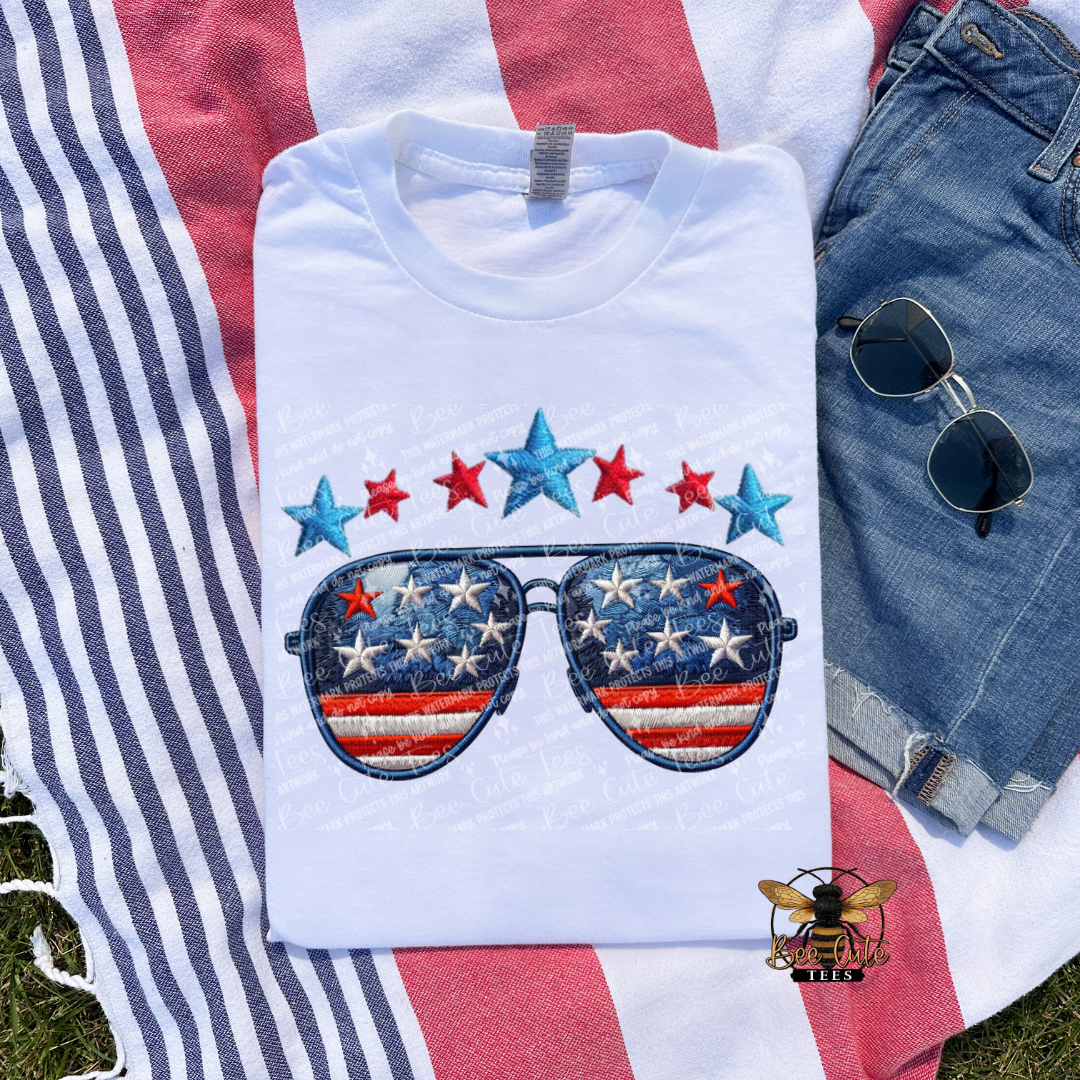 White t-shirt with star and stripe design on sunglasses, placed on a red and white striped surface with jeans and sunglasses.