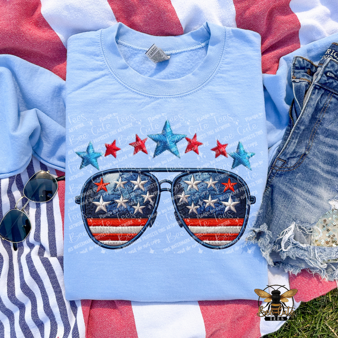 Light blue t-shirt with American flag sunglasses design on a red, white, and blue background.