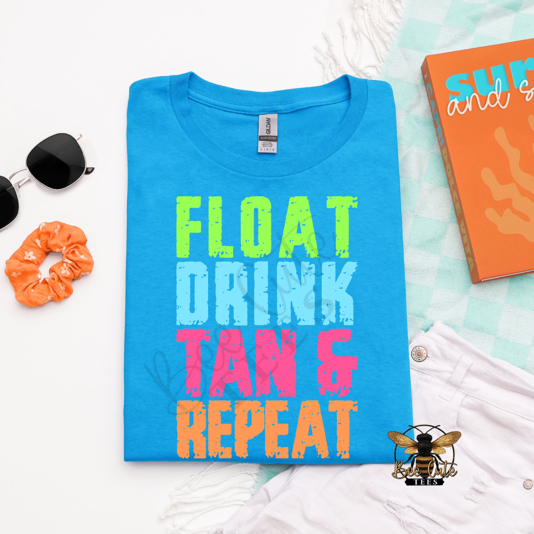 Folded bright blue graphic tee with colorful distressed text that reads “Float Drink Tan & Repeat,” styled with sunglasses, an orange scrunchie, white shorts, and beach-inspired accessories.
