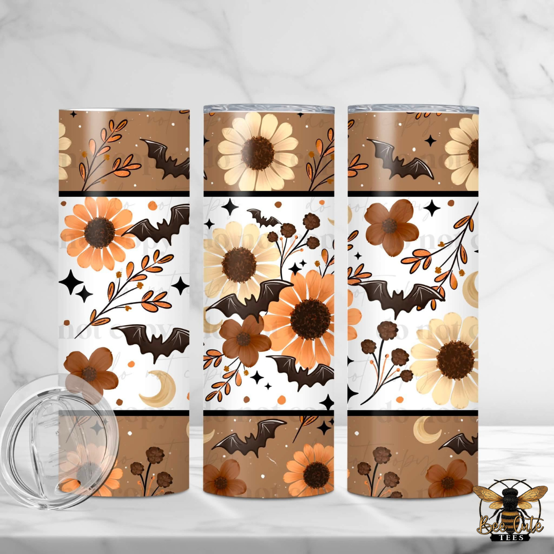 Three tall insulated tumblers with a Halloween-themed wrap design featuring orange and cream sunflowers, brown bats, and autumn leaves on a white and tan pattern. A clear plastic lid is shown in the lower left on a white marble surface.