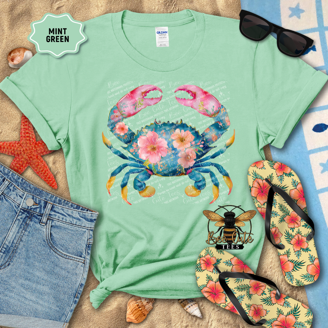 Mint green t-shirt with a colorful crab design on a beach-themed background with sunglasses, flip-flops, and starfish.
